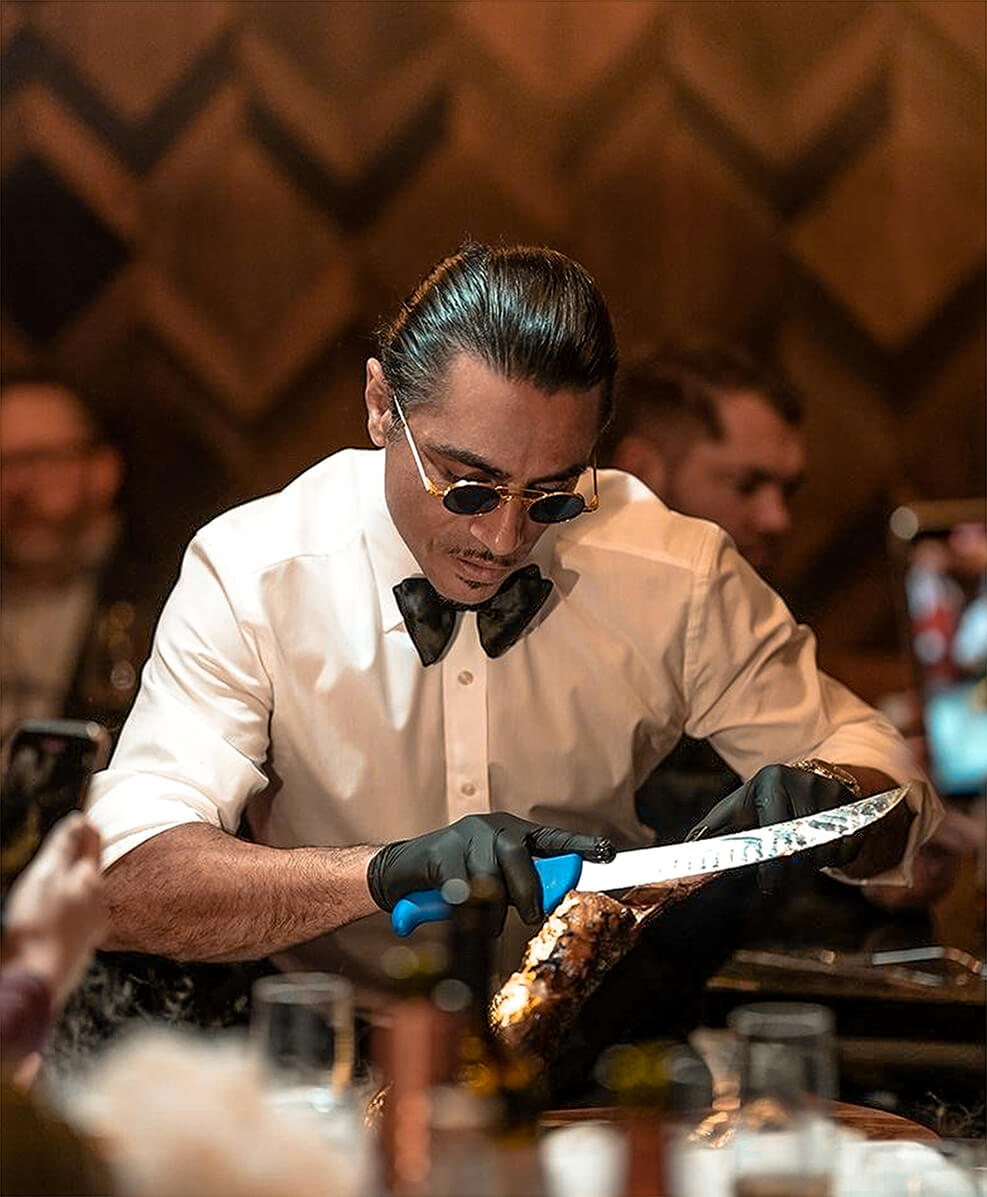 Şef Nusret Gökçe, beyaz tişörtü ve koyu gözlükleriyle ikonik pozunda, imzası olan tuz dökme hareketiyle bir bifteği lezzetlendiriyor